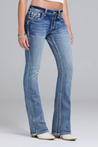 A model wearing a pair of medium-wash blue bootcut jeans with contrast stitching, paired with white heeled boots against a light gray studio background.