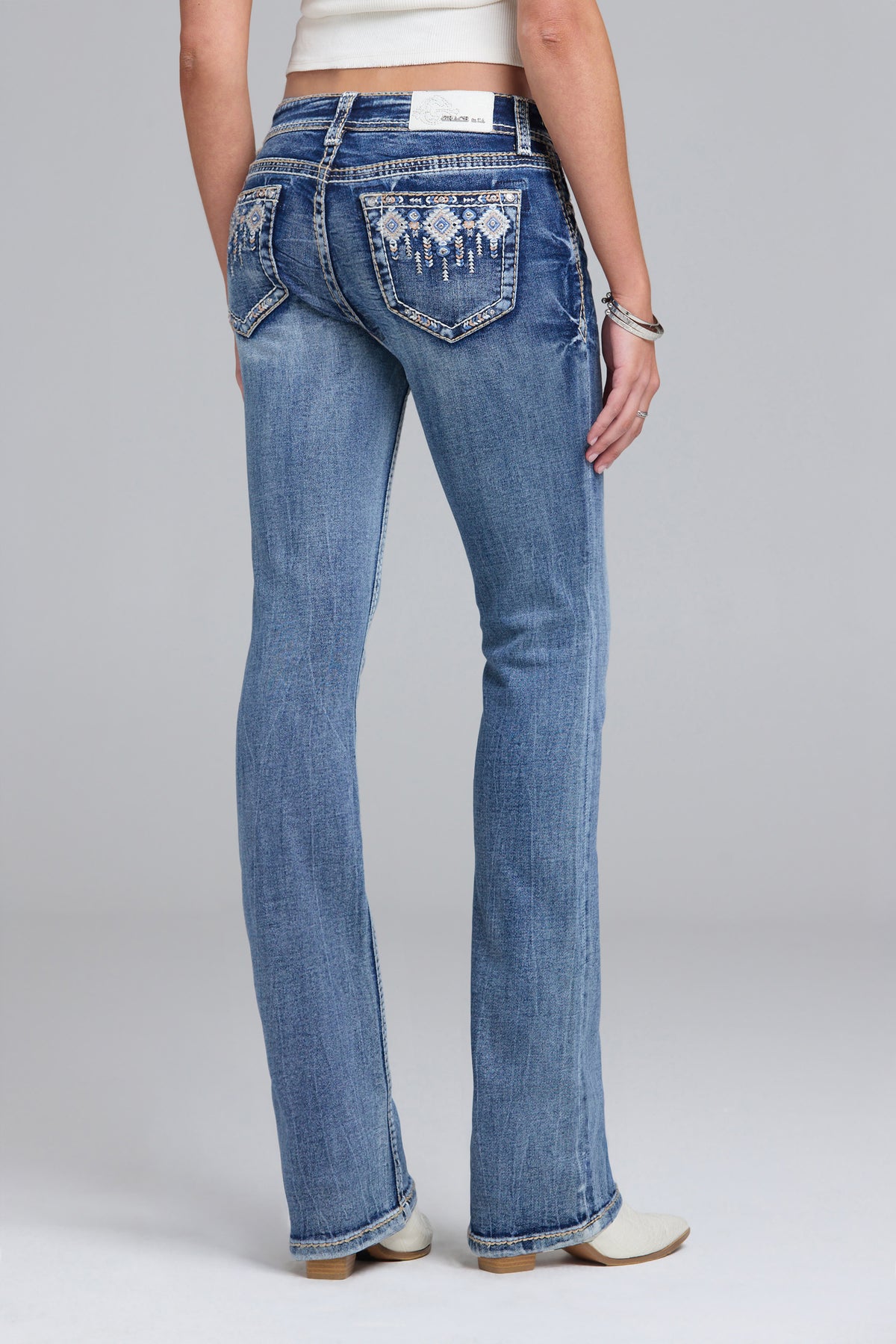 Rear view of a model wearing blue denim bootcut jeans with decorative embroidered detailing on the back pockets, paired with a white crop top and white shoes, against a light gray studio background.