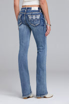 Rear view of a model wearing blue denim bootcut jeans with decorative embroidered detailing on the back pockets, paired with a white crop top and white shoes, against a light gray studio background.